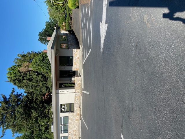 A one storey commercial building with white walls, a mostly flat gray roof, and a foundation of yellow, gray and white stone is situated in a parking lot paved in gray concrete or asphalt, with marked parking spaces.